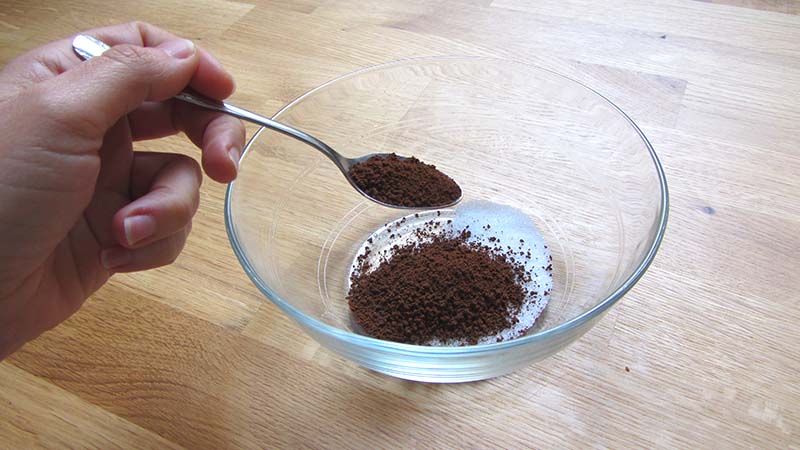 Una mano echa una cucharadita de café soluble en un cuenco transparente con azúcar, sobre una encimera de madera