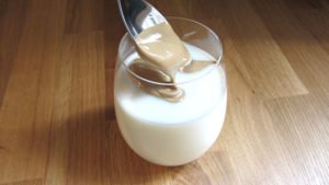 Una cuchara echa la crema de Café Dalgona dentro de un vaso de cristal de leche, sobre encimera de madera